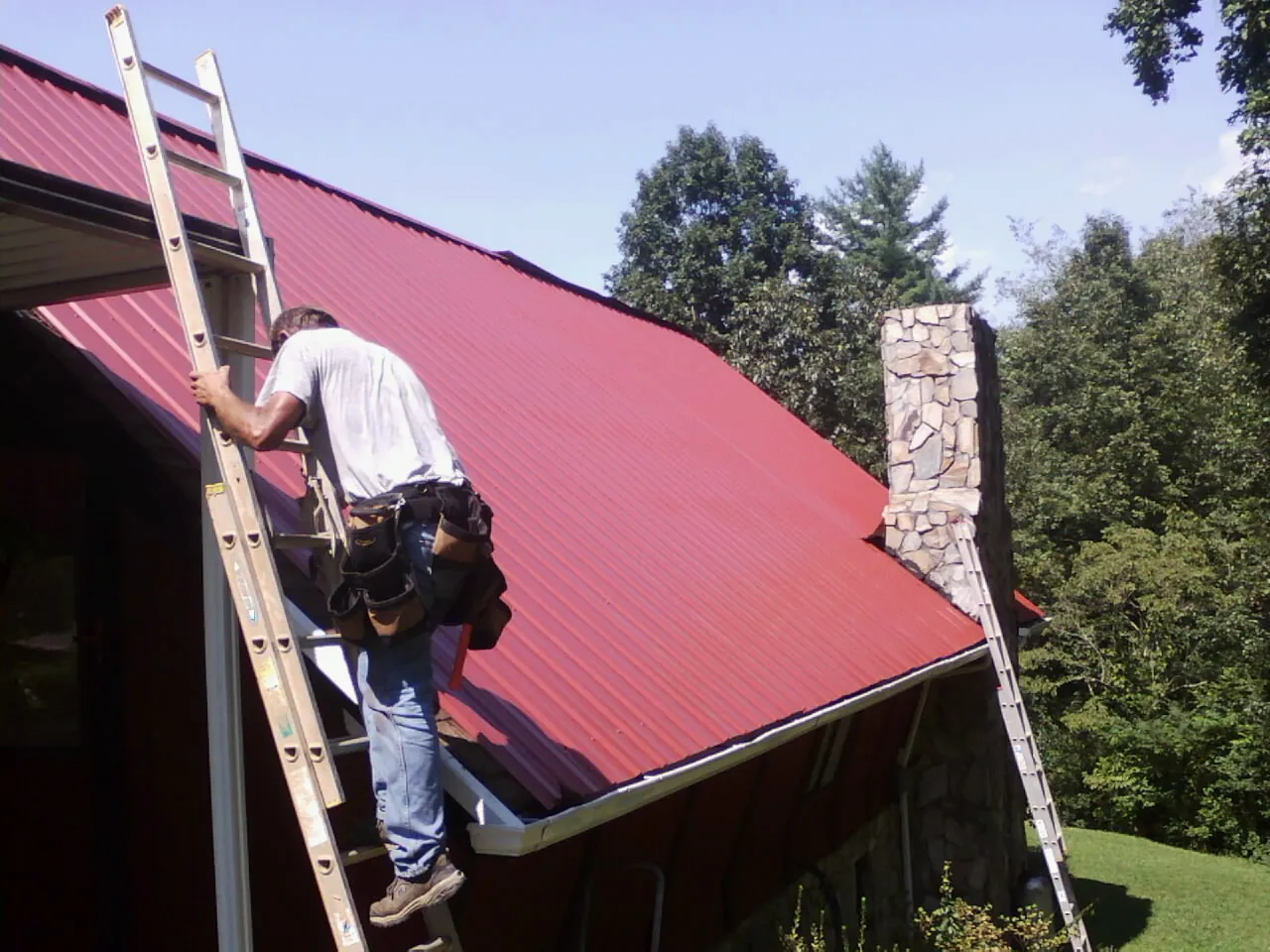 Roofer on a ladder beside a red metal roof and stone chimney — Soffit & Fascia Repair by Roofing Brothers in Chesterville